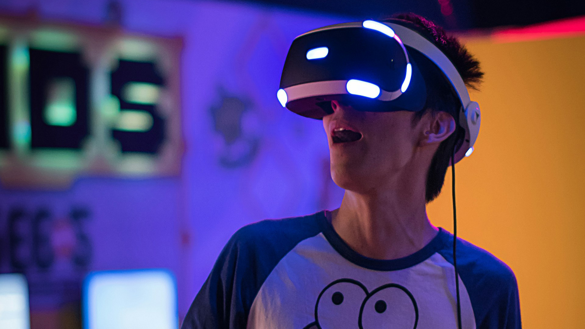 Un jeune homme porte un casque de réalité virtuelle avec des lumières bleues allumées, bouche entrouverte en signe d’étonnement, dans une salle aux lumières colorées
